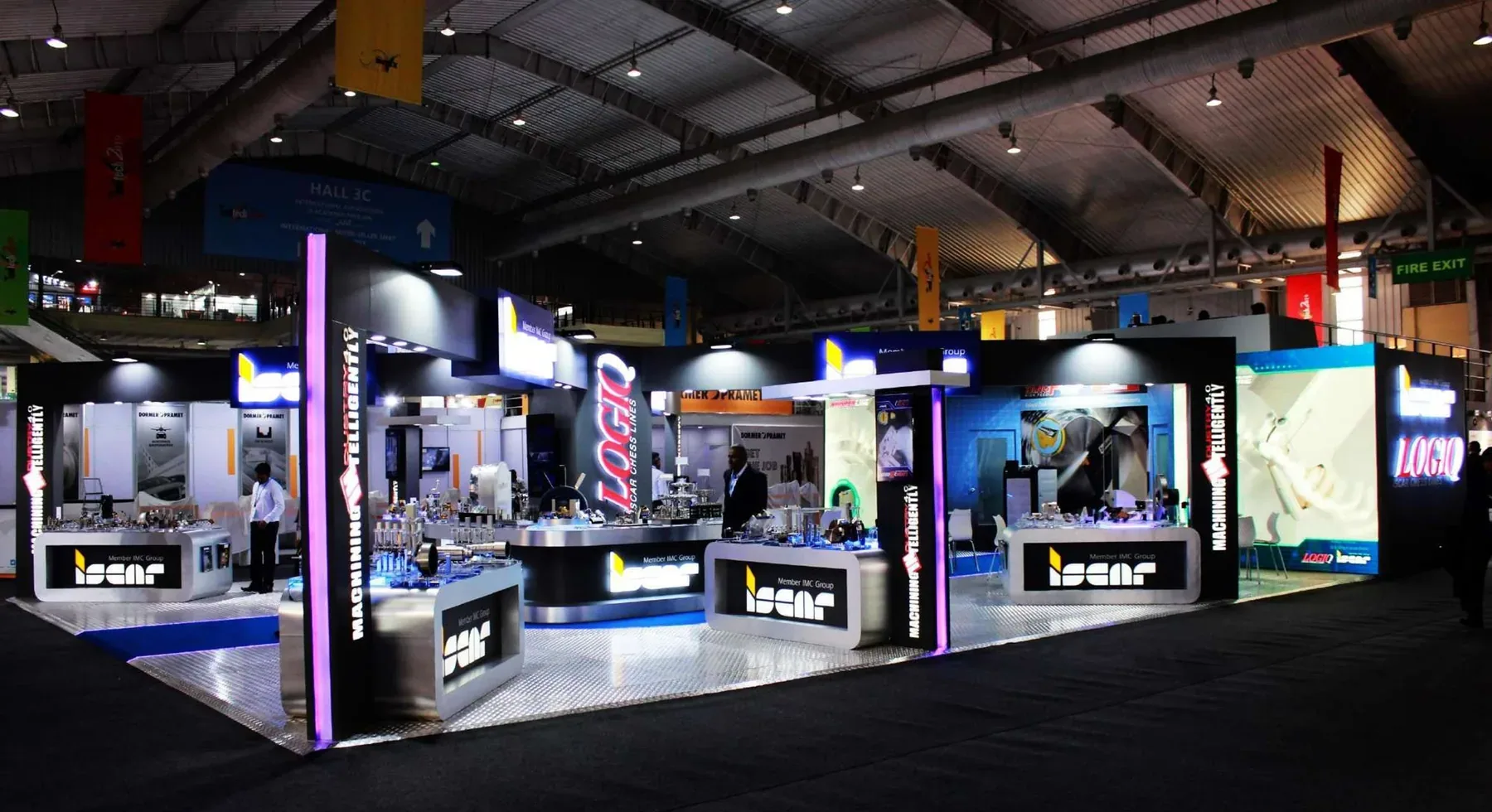 A large dark ISCAR exhibition space with backlit magenta columns, illuminated product counters and a checkerplate floor under an industrial hall roof