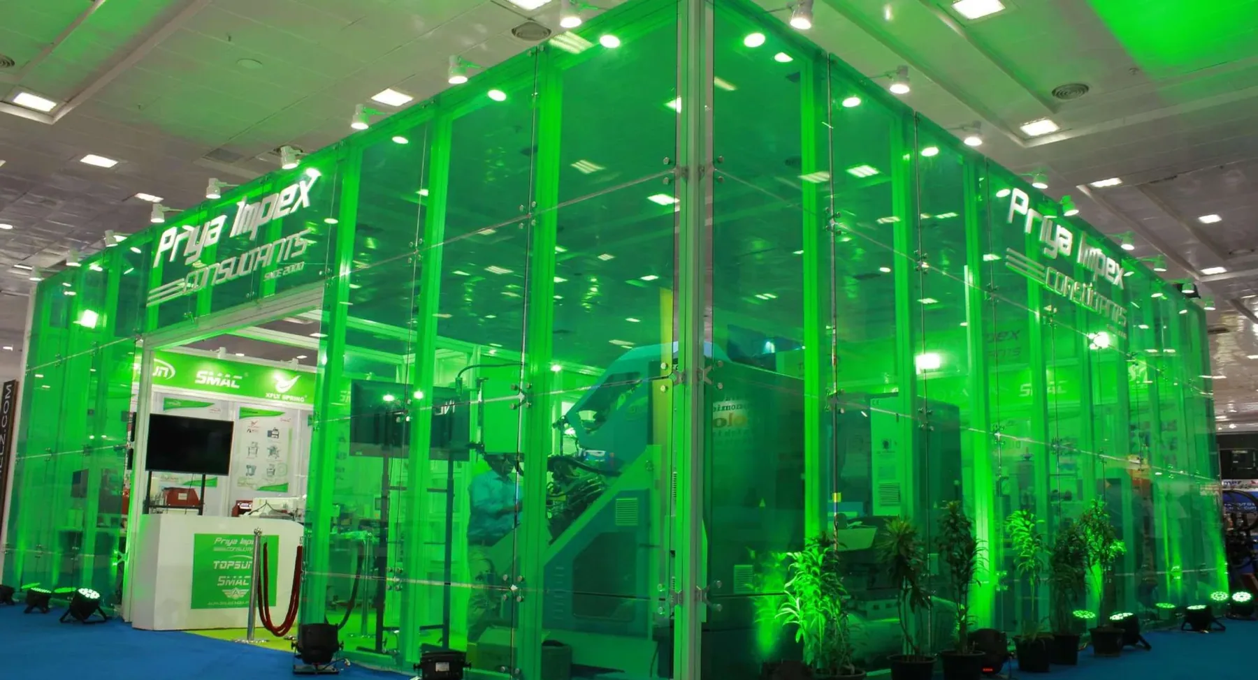 A double-height glass-walled exhibition pavilion uplit in saturated green, with the client name printed across two elevations