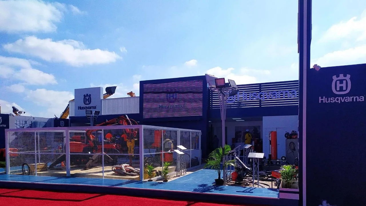 An outdoor exhibition pavilion under open sky, with a mesh-enclosed machinery compound in front of a navy louvred façade