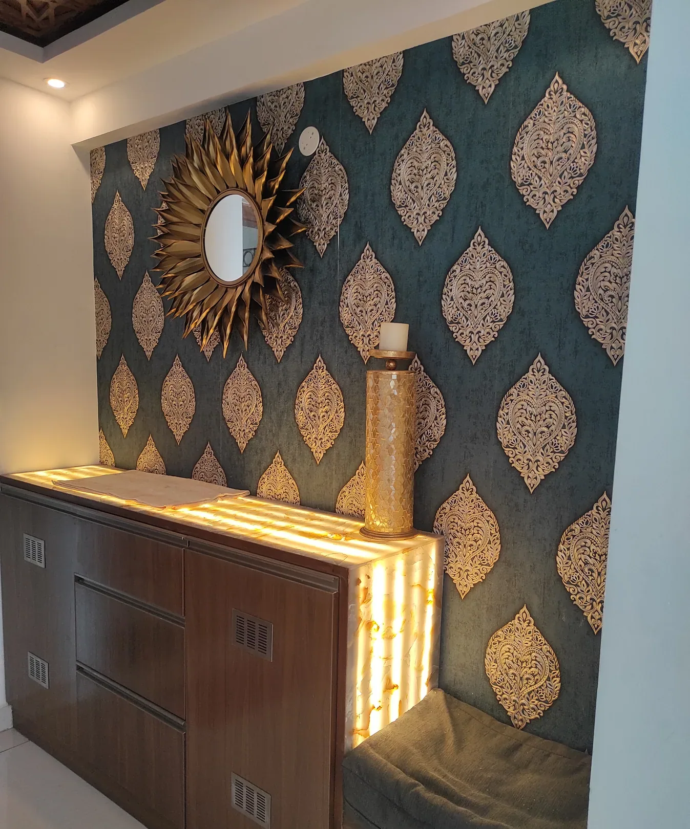 A residential entrance console in dark timber with a backlit stone-look panel, set against teal and gold patterned wallpaper below a sunburst mirror